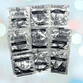 A grid of eight silver wrappers with black text, showcasing the bCondoms brand logo, the label "LARGE", and the phrase "SUPPORT BLACK BUSINESSES." The wrappers contain b Condoms Platinum Large Condoms, which are odor-free. They are set against a softly blurred background adorned with circular bokeh lights in various pastel colors.
